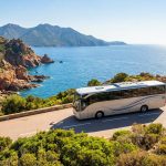 Un car blanc roule sur une route côtière sinueuse en Corse, avec la mer Méditerranée bleue et des montagnes rocheuses verdoyantes sous un ciel clair.
