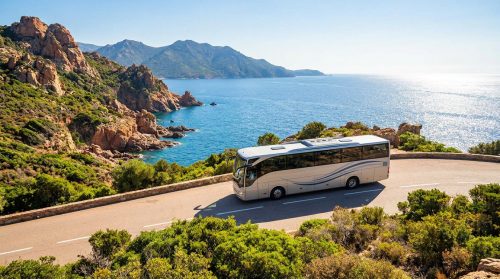 Un car blanc roule sur une route côtière sinueuse en Corse, avec la mer Méditerranée bleue et des montagnes rocheuses verdoyantes sous un ciel clair.