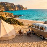 Vue aérienne d'un camping sur une plage sarde. Une tente beige, un van vintage et deux personnes en chaises face à la mer turquoise et aux falaises.