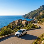 Voiture blanche sur route sinueuse le long de la côte du Cap Corse, avec un village traditionnel en pierre et la mer Méditerranée. Jour ensoleillé.