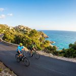 Deux cyclistes sur une route côtière sinueuse en Corse, avec un village perché et la mer Méditerranée scintillante au loin.