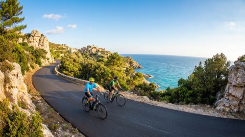 Deux cyclistes sur une route côtière sinueuse en Corse, avec un village perché et la mer Méditerranée scintillante au loin.