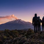 Groupe de randonneurs de dos observant l'Etna enneigé et fumant au lever du soleil sur un paysage volcanique rocailleux.