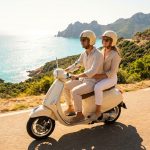 Couple en scooter beige sur une route côtière ensoleillée. La mer turquoise et les collines verdoyantes de Corse sont en arrière-plan.