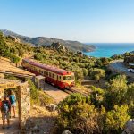 Groupe de voyageurs à un arrêt de bus/train en Corse, avec un bus sur une route côtière et un train à quai, face à la mer turquoise.
