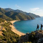 Deux randonneurs contemplent un paysage côtier corse avec montagnes, route sinueuse, plage de sable et mer turquoise.