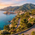 Route côtière sinueuse surplombant la mer turquoise et un village portuaire méditerranéen en Corse au coucher du soleil.