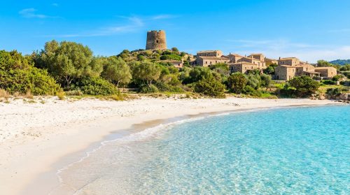 Plage de sable blanc aux eaux turquoise avec village de maisons en pierre et tour historique en arrière-plan, sous un ciel bleu.