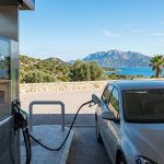 Voiture grise faisant le plein à une station-service. L'écran affiche 78.50€ pour 45.5 litres. En fond, paysage côtier corse avec mer et montagnes.