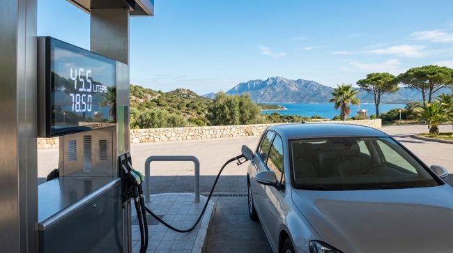 Voiture grise faisant le plein à une station-service. L'écran affiche 78.50€ pour 45.5 litres. En fond, paysage côtier corse avec mer et montagnes.