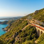 Un train rouge et blanc, le Trinighellu, traverse un viaduc en pierre le long d'une côte rocheuse et verdoyante de Corse, avec la mer turquoise à gauche.