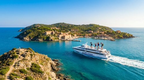 Ferry Moby naviguant vers un village côtier méditerranéen sur une île verdoyante, sous un ciel bleu clair.