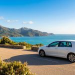 Voiture blanche garée sur une route côtière en Corse avec la mer turquoise et les montagnes verdoyantes en arrière-plan.