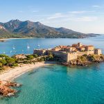 Vue aérienne de la citadelle de Porto-Vecchio en Corse, entourée d'une mer turquoise et de montagnes en arrière-plan.