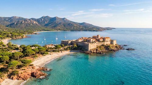 Vue aérienne de la citadelle de Porto-Vecchio en Corse, entourée d'une mer turquoise et de montagnes en arrière-plan.