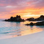 Découvrez les plus belles plages de Corse pour vos vacances Plage de sable blanc avec des rochers, baignée par les couleurs orangées d'un coucher de soleil corse.