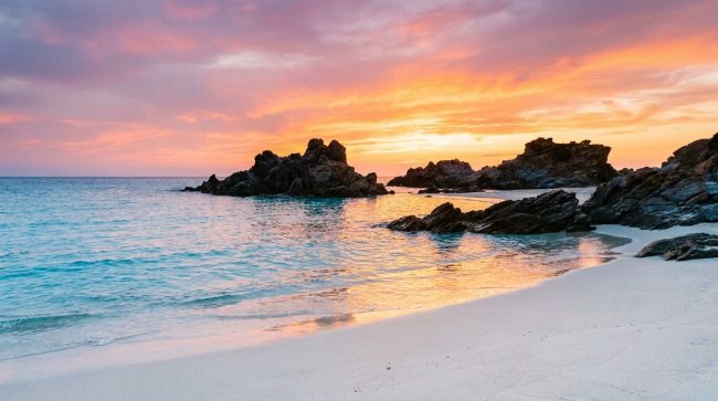 Plage de sable blanc avec des rochers, baignée par les couleurs orangées d'un coucher de soleil corse.