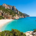 Que faire en Sardaigne : les lieux clés de votre voyage Une crique isolée avec une plage de sable blanc et des falaises rocheuses surplombant une mer turquoise en Sardaigne.