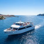Un ferry blanc navigue sur une mer bleue, avec des passagers à bord et la côte méditerranéenne en arrière-plan.