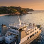 Ferry transportant des passagers et un camping-car naviguant le long des côtes rocheuses de Corse au coucher du soleil.