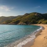 Une personne marche sur une plage de sable en Corse avec une mer turquoise et des collines verdoyantes en arrière-plan.
