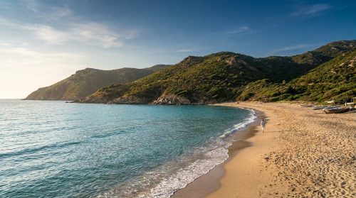 Une personne marche sur une plage de sable en Corse avec une mer turquoise et des collines verdoyantes en arrière-plan.