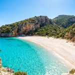 Plage paradisiaque en Sardaigne aux eaux turquoise cristallines bordée de falaises rocheuses et de végétation luxuriante.