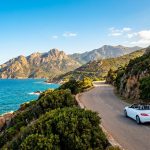 Une voiture décapotable blanche roulant sur une route côtière sinueuse surplombant la mer Méditerranée en Corse.