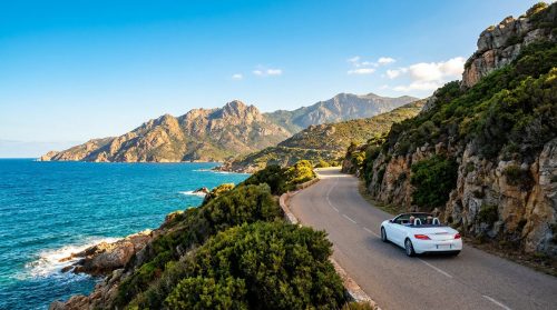 Une voiture décapotable blanche roulant sur une route côtière sinueuse surplombant la mer Méditerranée en Corse.