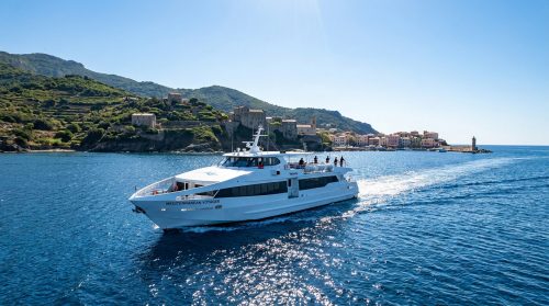 Le ferry Mediterranean Voyager naviguant en mer Méditerranée devant un village côtier Corse sous un ciel bleu.