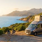 Un couple profite de la vue sur la mer en Corse, assis près de leur van aménagé avec toit relevable au coucher du soleil.